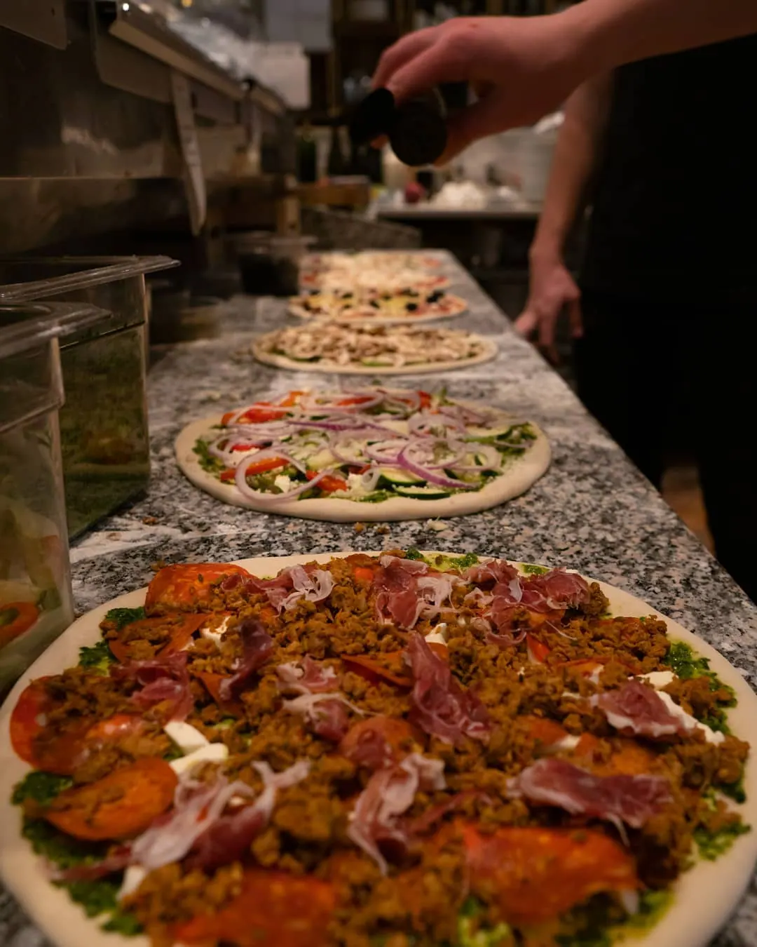 Pizzas lined up on a granite counter while a cook finishes them.
