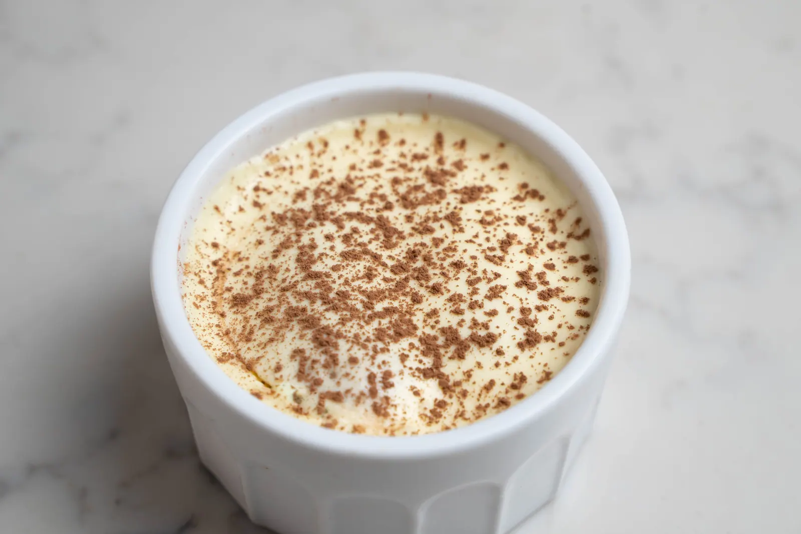 Tiramisu dusted with cocoa in a white ramekin on a marble counter.